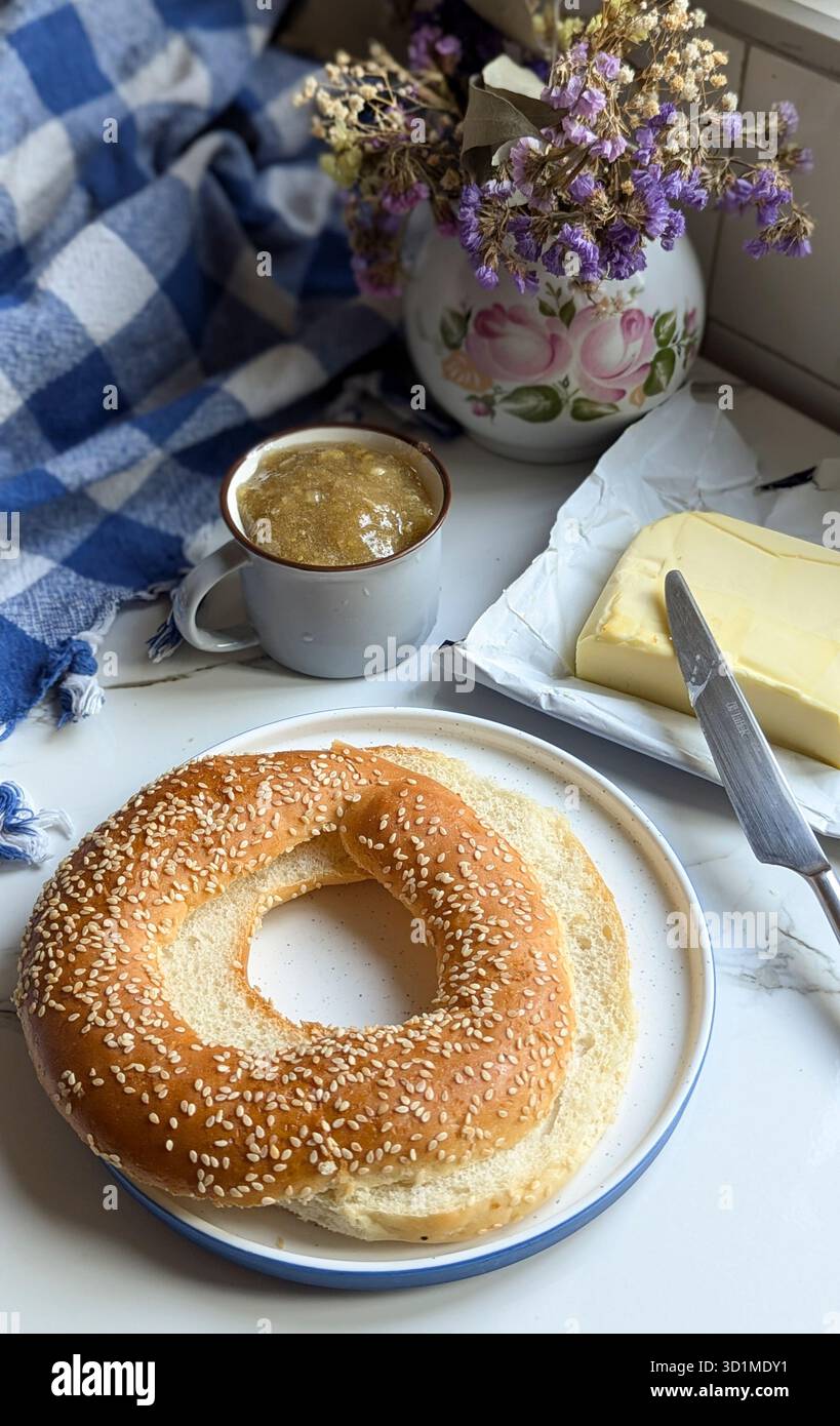 Bagel de sésame frais servi avec du beurre et une tasse de café sur une table de petit déjeuner confortable près de la fenêtre. Scène matinale rustique avec lumière naturelle, fait maison f - Image de stock capturée avec un smartphone
