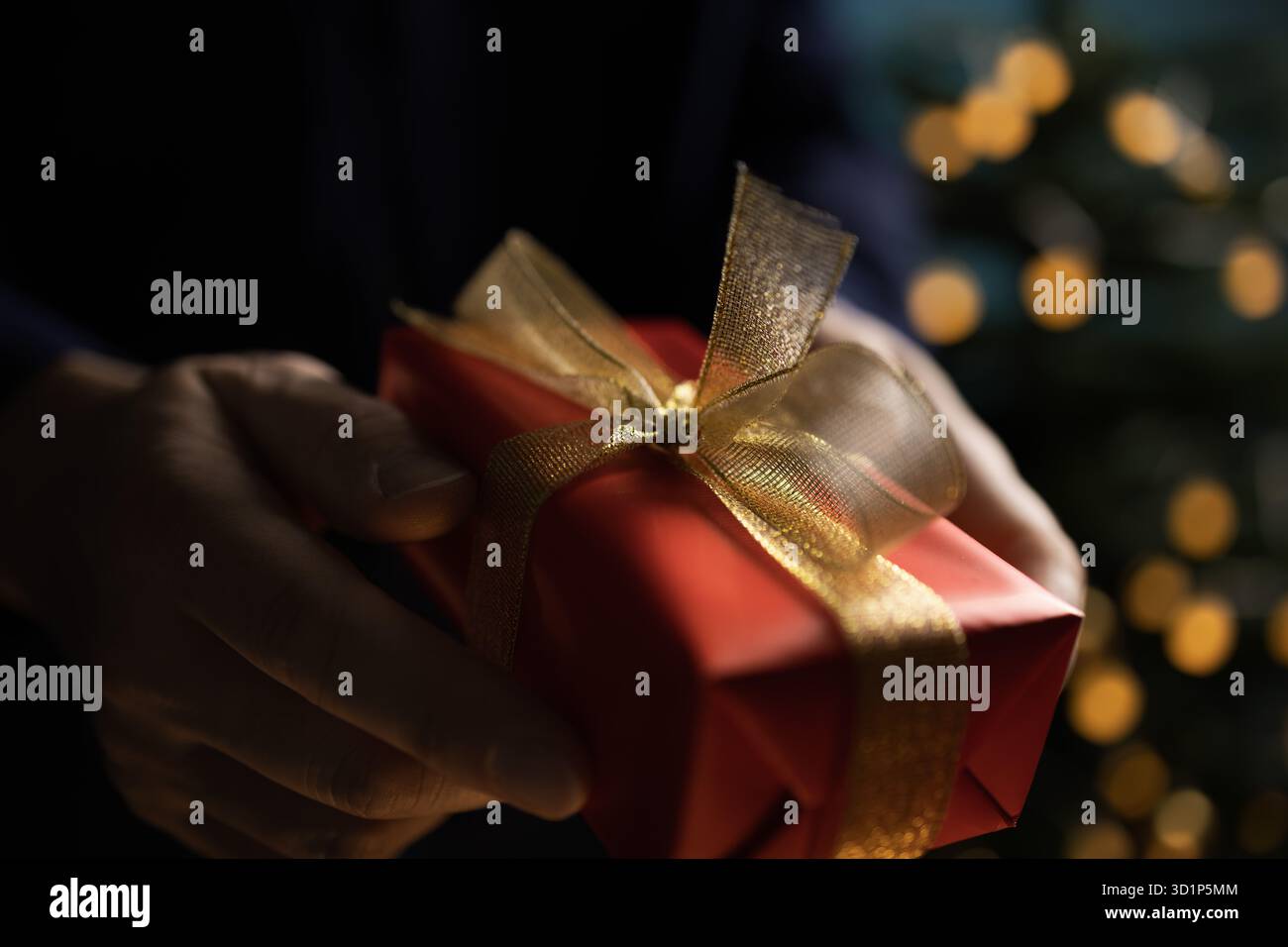 Homme tenant un cadeau de Noël emballé près d'un arbre de Noël décoré avec des lumières festives et une atmosphère de vacances confortable Banque D'Images