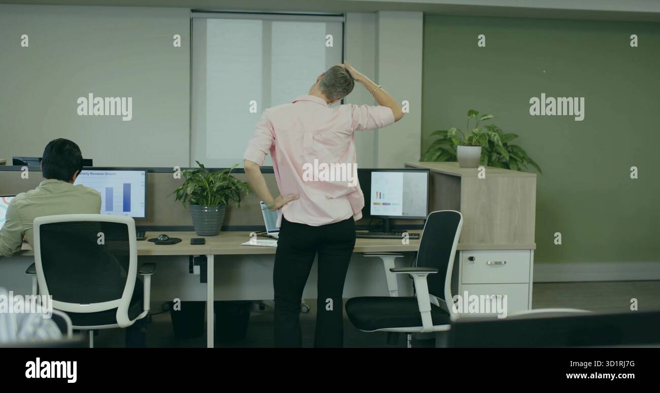 Travailleuse de bureau portant une chemise rose tenant la tête dans le bureau, avec des moniteurs et des plantes en pot Banque D'Images
