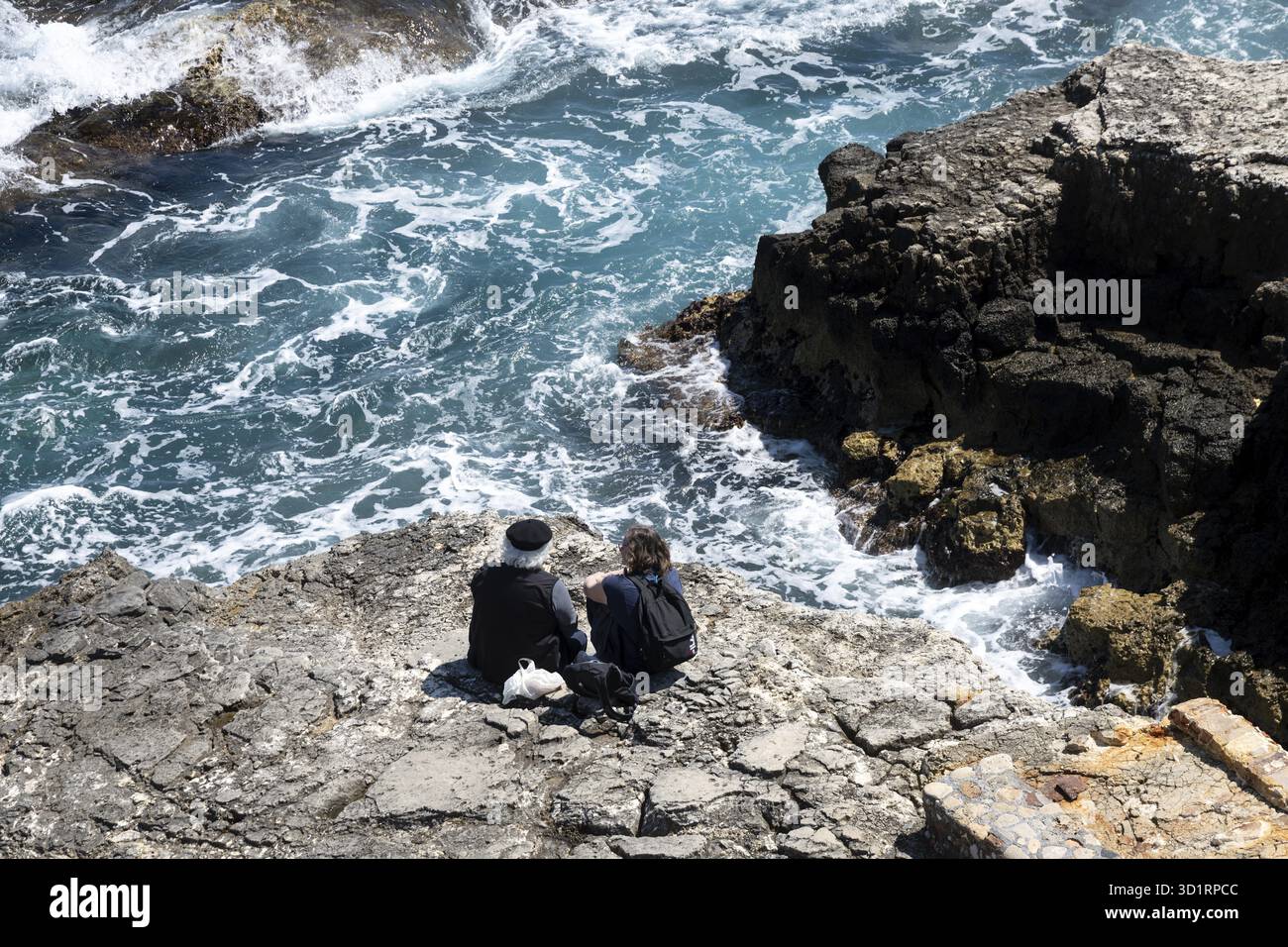 Syrakus, Sizilien - Italie - 04-09-2025 : deux personnes assises sur la rive rocheuse à Syracuse regardant la mer bleue avec des vagues blanches s'écraser contre la côte un Banque D'Images