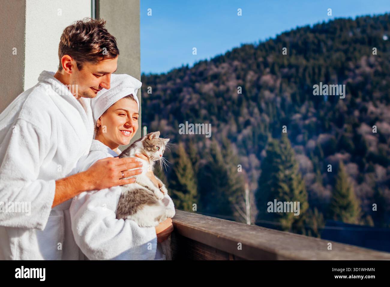 Jeune couple tenant chat sur le balcon de l'hôtel profitant du paysage de montagne d'hiver portant peignoir. Voyagez avec votre animal de compagnie. Homme et femme se relaxant en jouant avec Do Banque D'Images