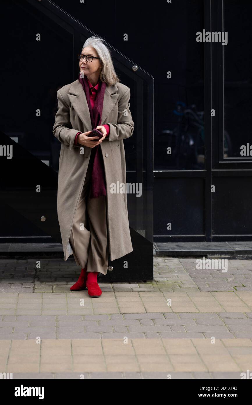 Femme âgée élégante aux cheveux gris habillée d'un long manteau et de chaussures rouges se tient gracieusement contre un mur noir moderne dans un ur Banque D'Images