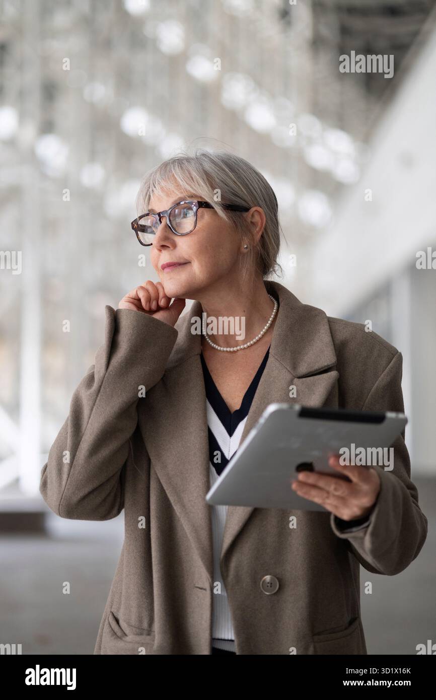 Femme d'affaires d'âge moyen considère soigneusement ses prochaines étapes tout en tenant une tablette dans un immeuble de bureaux moderne pendant la da Banque D'Images