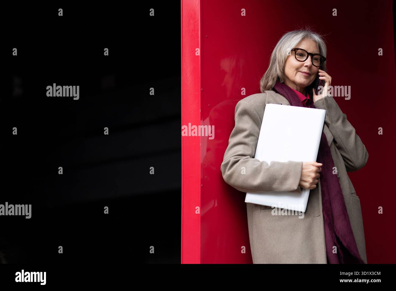 Femme d'affaires âgée dans un manteau élégant se tient en toute confiance, tenant un ordinateur portable tout en parlant sur son téléphone dans un settin urbain moderne Banque D'Images