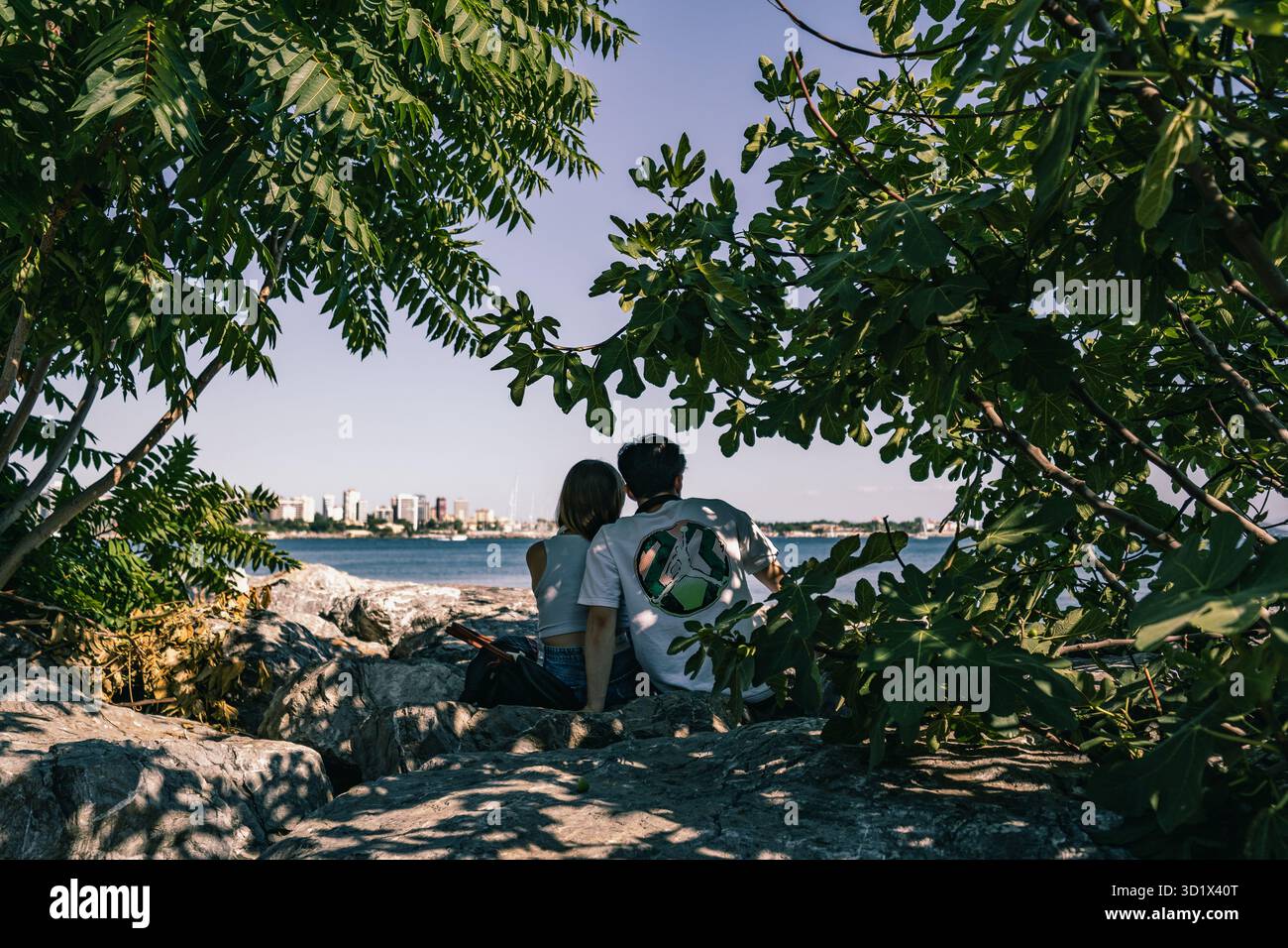 Couple assis au bord de la mer de Marmara sous les arbres par une journée ensoleillée à Istanbul, Türkiye, partageant un moment de calme. Banque D'Images