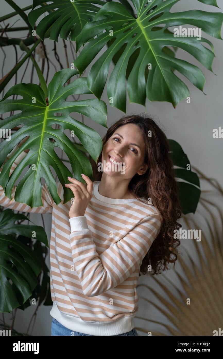 Jeune femme positive heureuse touchant la feuille de Monstera, passant du temps avec les plantes dans le jardin à la maison Banque D'Images