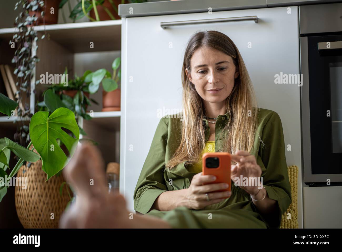 Femme souriante accroche dans un smartphone sur la pause des travaux ménagers, travail entouré de plantes dans la cuisine Banque D'Images