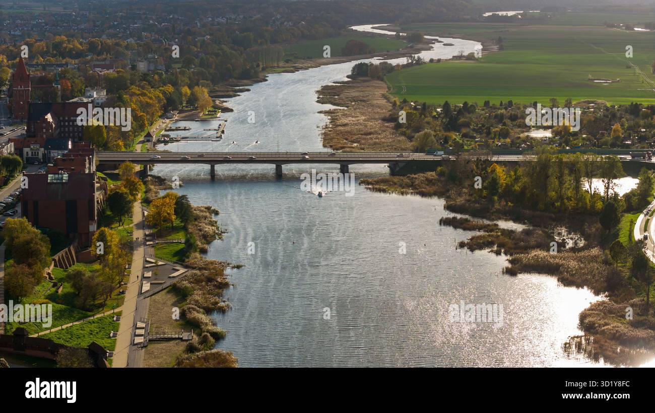 Vue par drone à grand angle de la rivière Nogat par Malbork, Pologne, avec un pont routier et un sillage de bateau à moteur, dépendances du château, roseaux, jardins et automne Banque D'Images