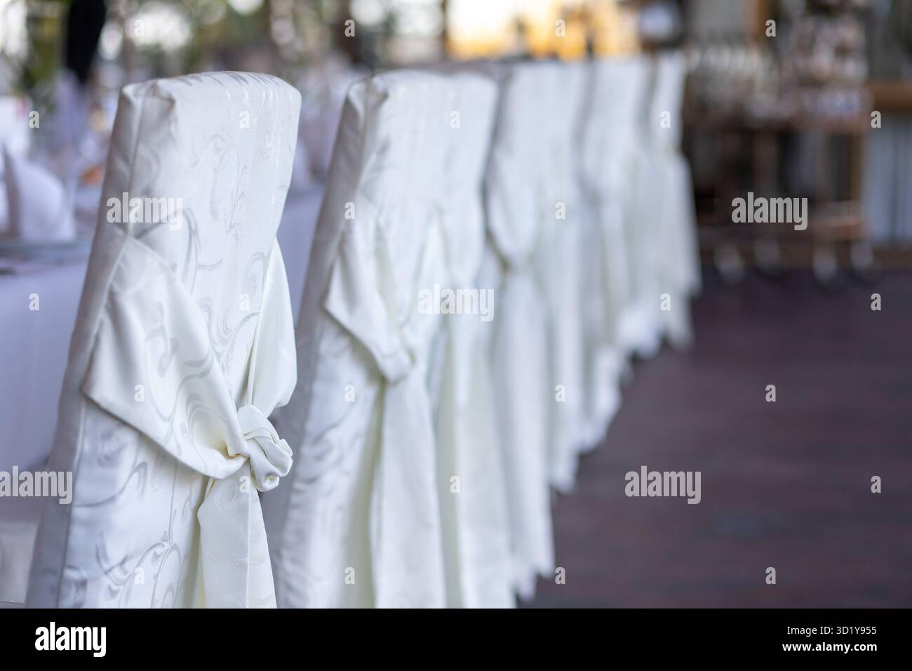 Une rangée de housses de chaises blanches élégantes est soigneusement agencée autour d'une longue table de banquet dans un lieu rustique. La lumière douce de l'après-midi souligne la configuration, creat Banque D'Images