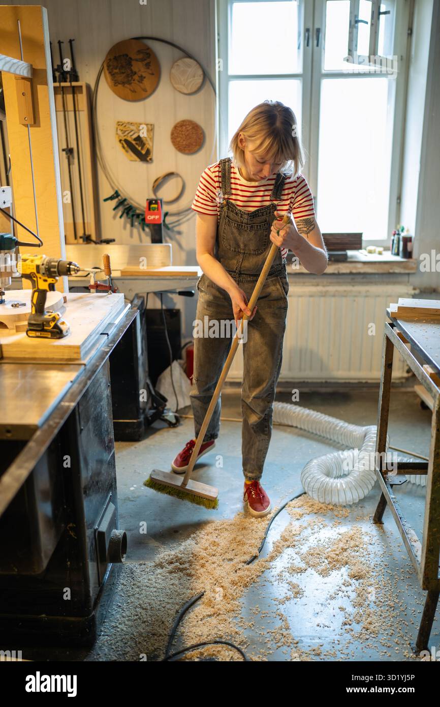 Femme charpentier nettoyant l'atelier après le travail. Handywoman balayer la sciure de bois et ventiler pour la santé Banque D'Images