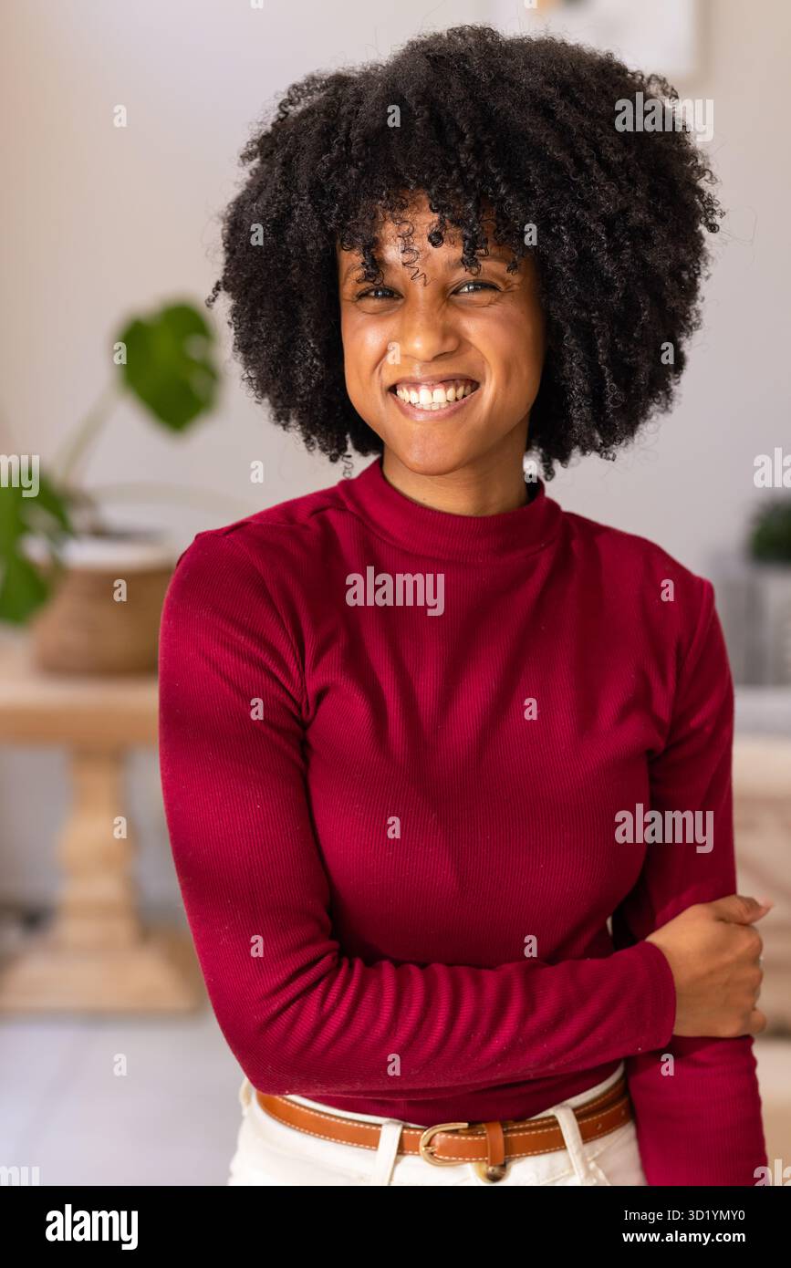 Femme afro-américaine posant portant un haut rouge, un pantalon blanc, une ceinture brune par console de maison avec plante Banque D'Images