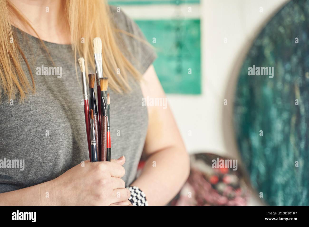 Artiste féminine caucasienne tenant des pinceaux en studio avec des œuvres d'art en arrière-plan. Banque D'Images