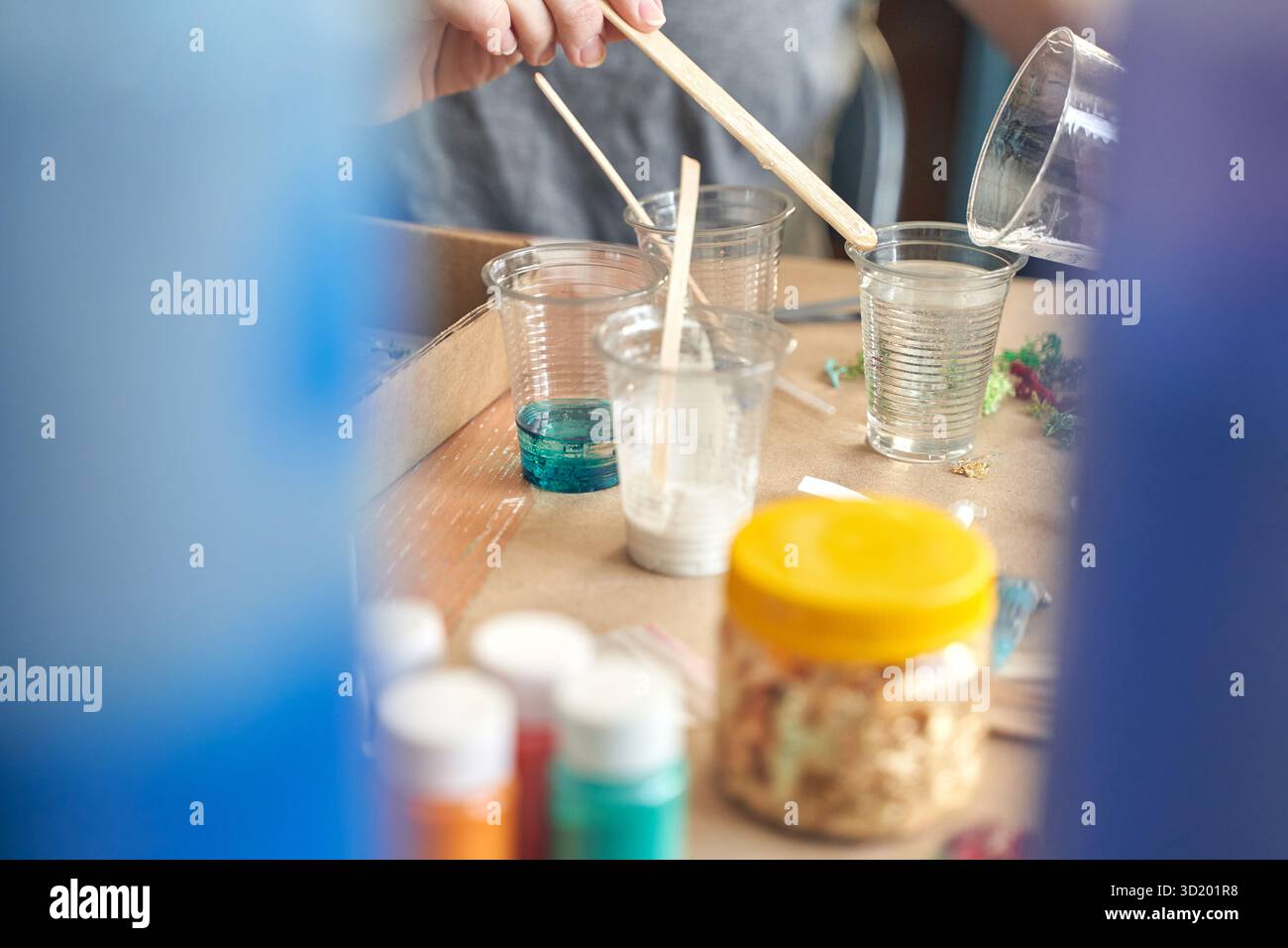 Mains versant de la résine dans des gobelets transparents pour le projet d'artisanat sur la table de travail avec de la peinture et des matériaux. Banque D'Images