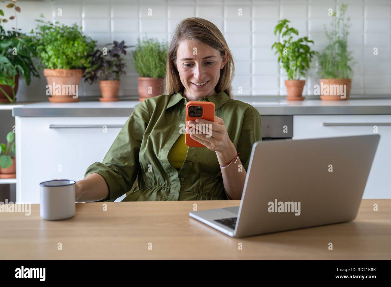 Femme joyeuse prenant une pause dans la cuisine, regardant sur smartphone chatter, le web défilant tenant le thé de tasse Banque D'Images