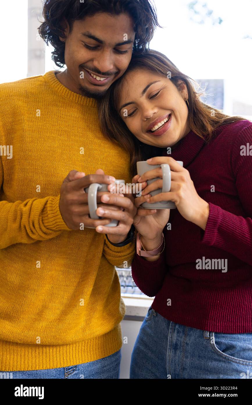 Couple indien penchant les têtes ensemble dans des chandails tricotés tenant des tasses en céramique par fenêtre à la maison Banque D'Images