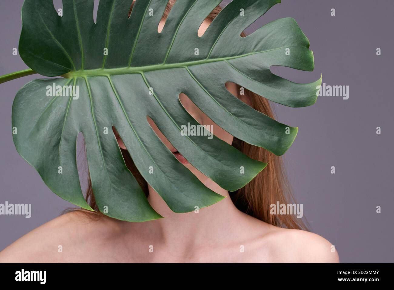 Mystérieuse jeune femme caucasienne partiellement cachée derrière une grande feuille de monstera Banque D'Images