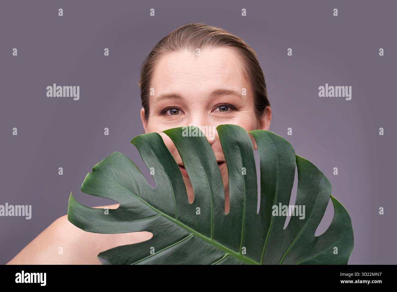 Souriante femelle adulte caucasienne jetant un coup d'œil de la feuille de monstera contre le gris Banque D'Images