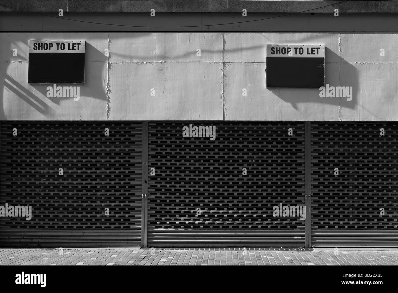 Boutique fermée avec volets métalliques et deux panneaux « Shop to Let » ci-dessus, capturés en noir et blanc, symbolisant le déclin urbain et l’espace commercial vacant. Banque D'Images