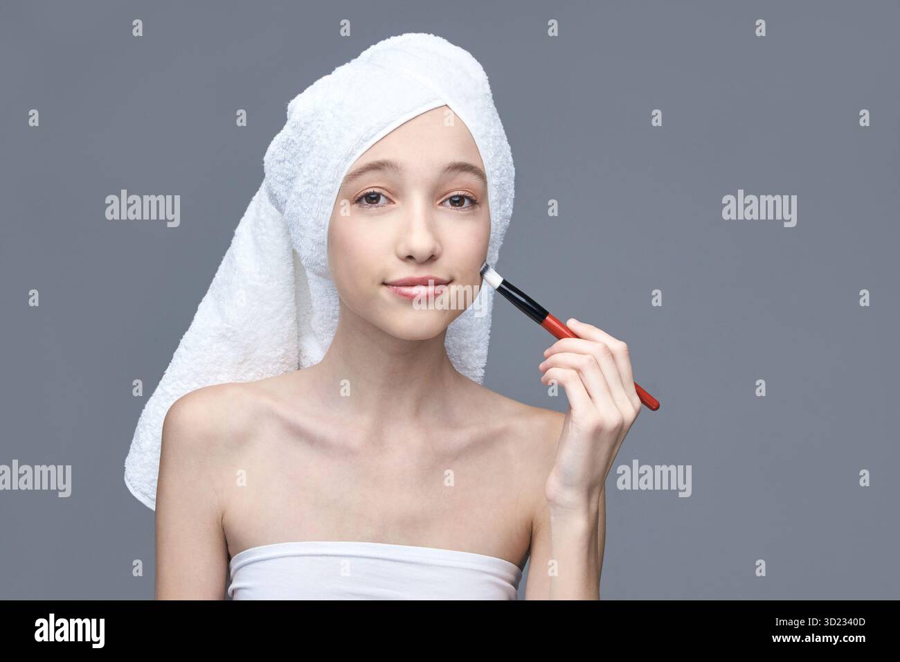 Jeune femme caucasienne avec des cheveux enveloppés de serviette appliquant le maquillage contre le gris Banque D'Images