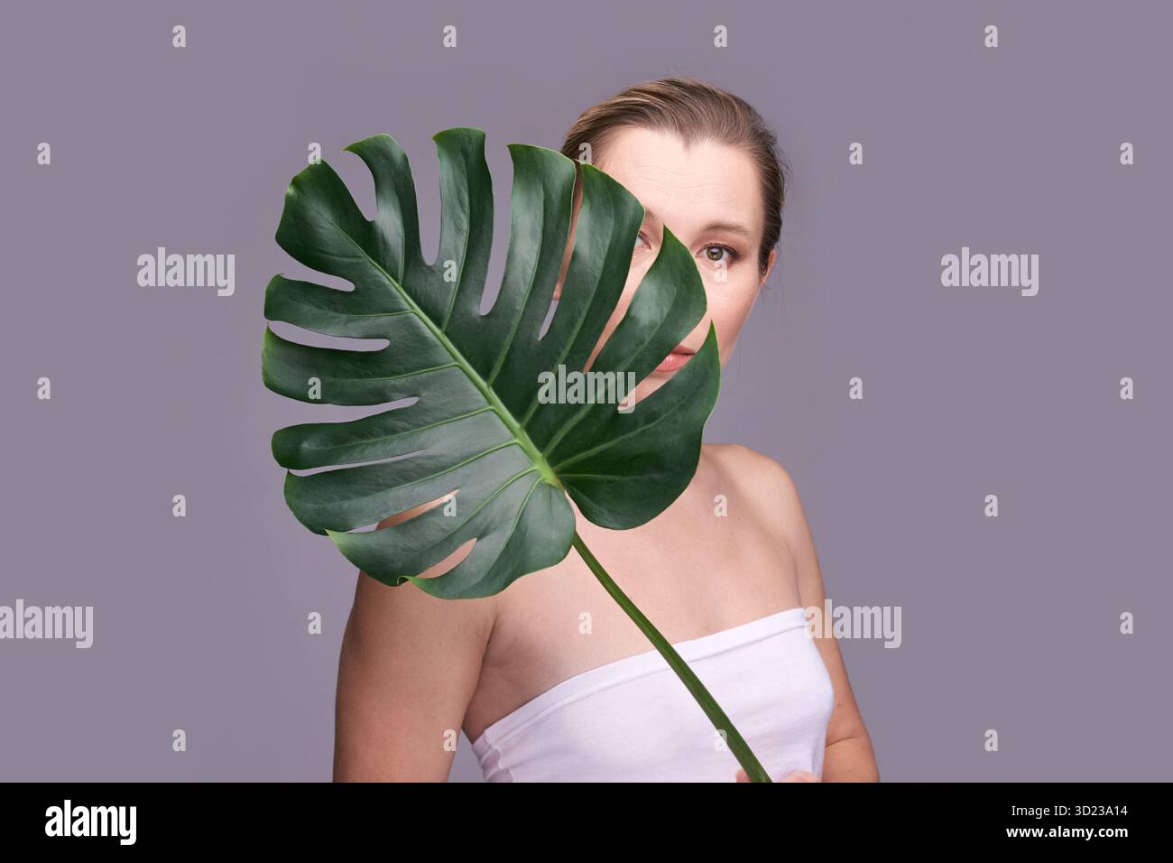 Femelle adulte caucasienne partiellement cachée derrière une grande feuille de monstera sur gris Banque D'Images