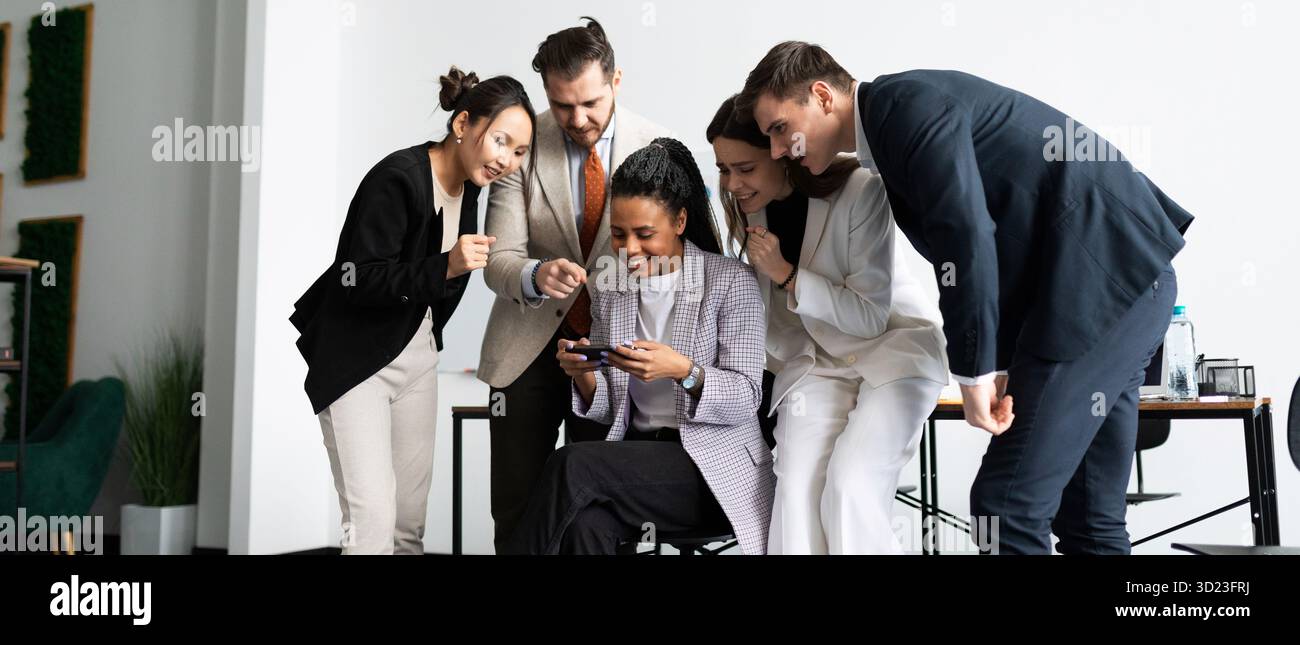 Une équipe de collègues au bureau regarde avec intérêt comment un employé joue sur un téléphone mobile Banque D'Images