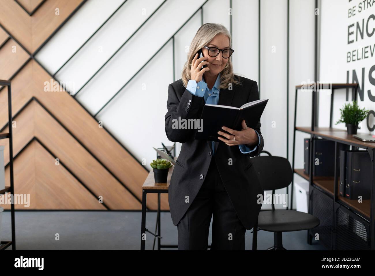 Femme gestionnaire de 50 ans dans un bureau stérile parle sur un téléphone portable avec un journal dans ses mains Banque D'Images