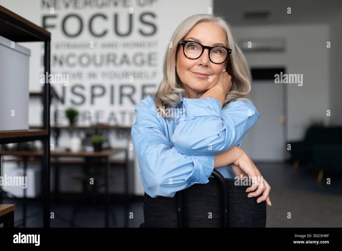 Femme d'affaires confiante avec des lunettes assis avec ses bras pliés confortablement sur le dos d'une chaise et regardant le camer Banque D'Images