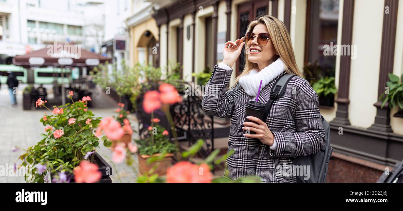Femme mature d'aspect européen caucasien redresse ses lunettes de soleil avec une tasse de café dans ses mains lors d'une promenade touristique en th Banque D'Images
