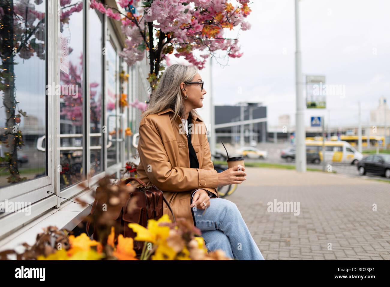 Une pensionnée élégante est assise sur un banc à côté d'un magasin avec un téléphone portable dans ses mains et regarde au loin Banque D'Images