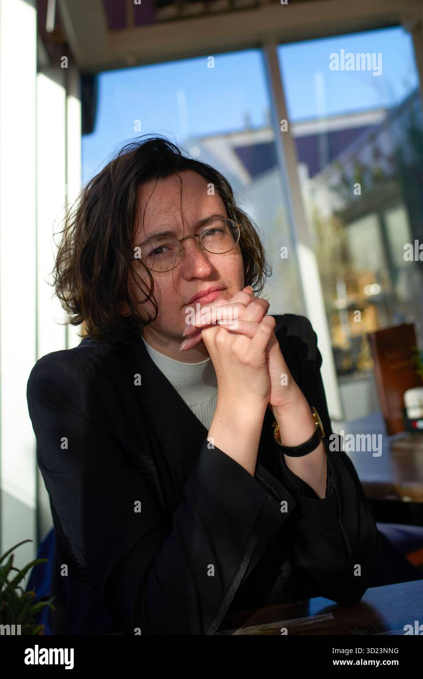 Jeune femme avec des lunettes assise au soleil près d'une fenêtre dans un café, les mains serrées et détendues. Blankenberge, Flandre occidentale, Belgique Banque D'Images