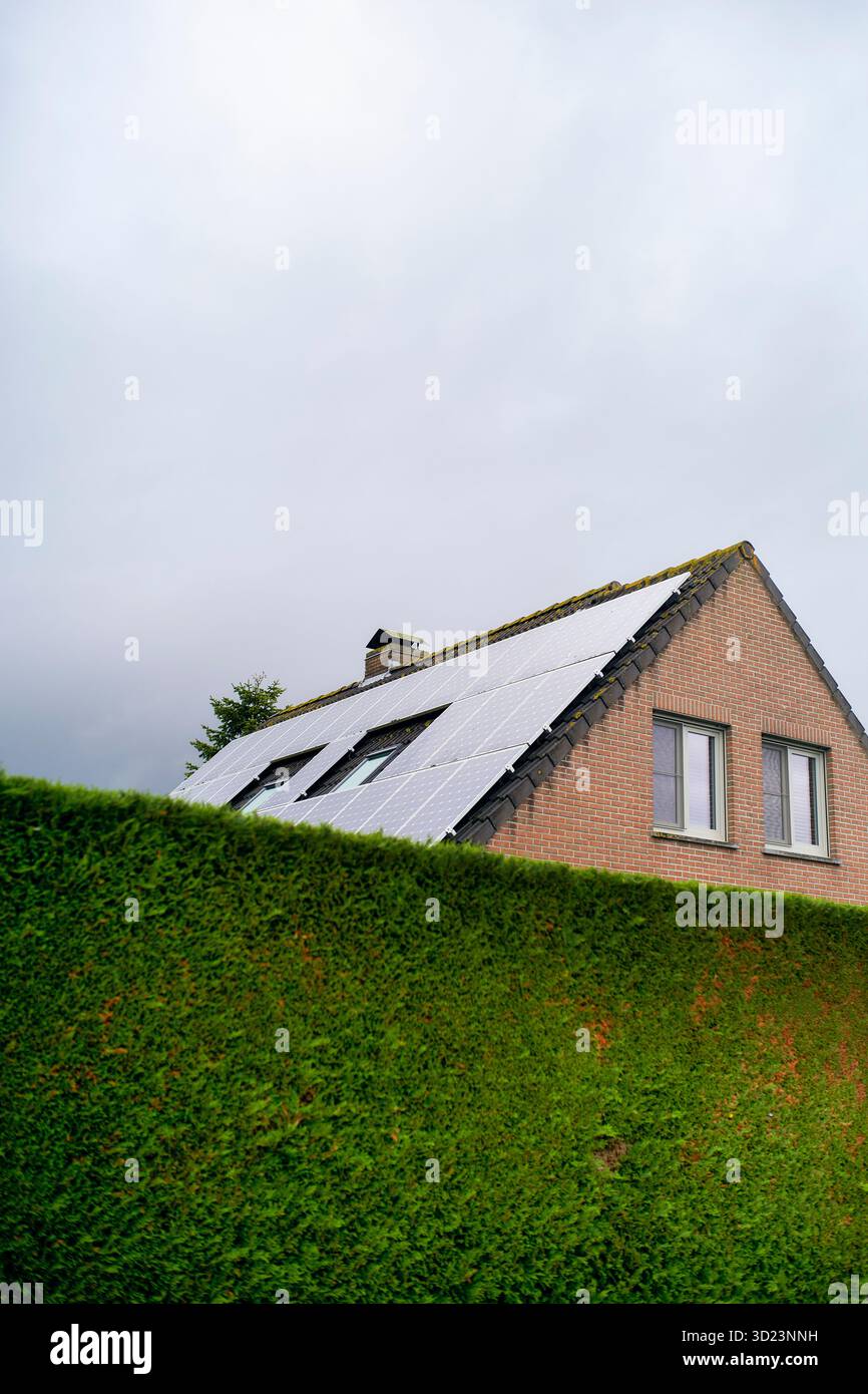 Maison en brique avec panneaux solaires sur le toit derrière une haie verte luxuriante sous le ciel nuageux. Blankenberge, Flandre occidentale, Belgique Banque D'Images