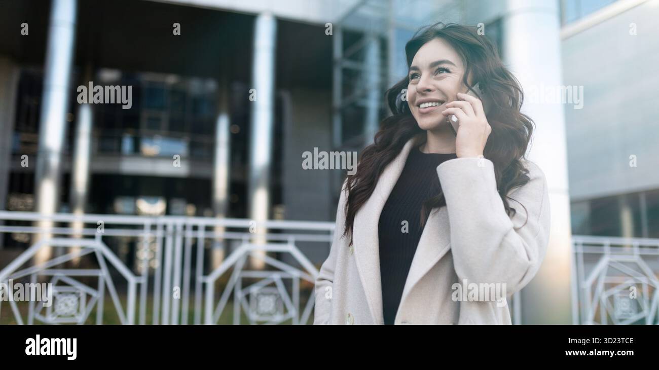 Jeune femme d'affaires confiante parlant sur le téléphone portable avec un grand sourire sur son visage à l'extérieur du bureau, WO forte et indépendante Banque D'Images