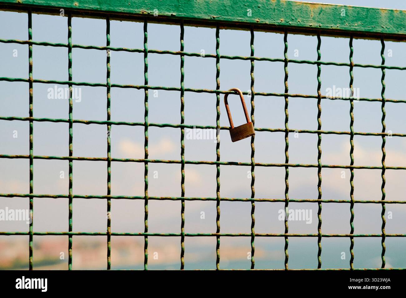 Un cadenas rouillé est accroché à une clôture en fil de fer vert avec une ligne d'horizon urbaine floue en arrière-plan. Banque D'Images