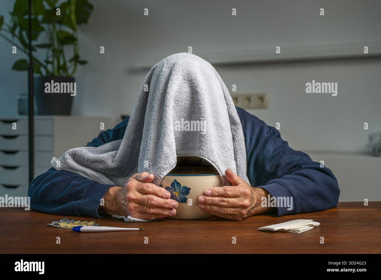 Homme âgé avec une serviette sur sa tête faisant inhalation de vapeur sur un bol avec de l'eau chaude à une table avec des pilules, un thermomètre de fièvre et des tissus, guérir Banque D'Images