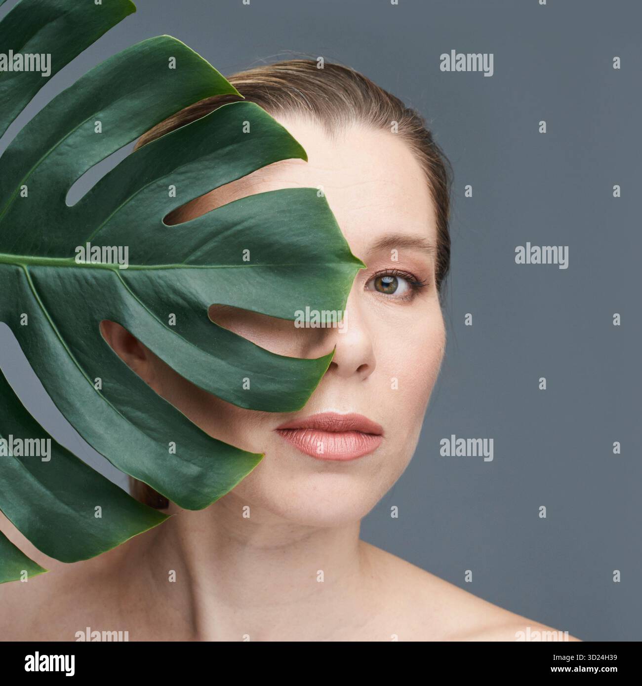 Jeune femelle caucasienne avec feuille de monstera sur fond gris exprimant naturel Banque D'Images
