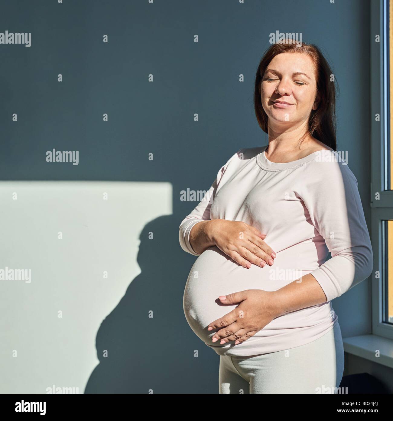 Femme caucasienne enceinte debout près de la fenêtre dans la lumière du soleil tenant le ventre Banque D'Images