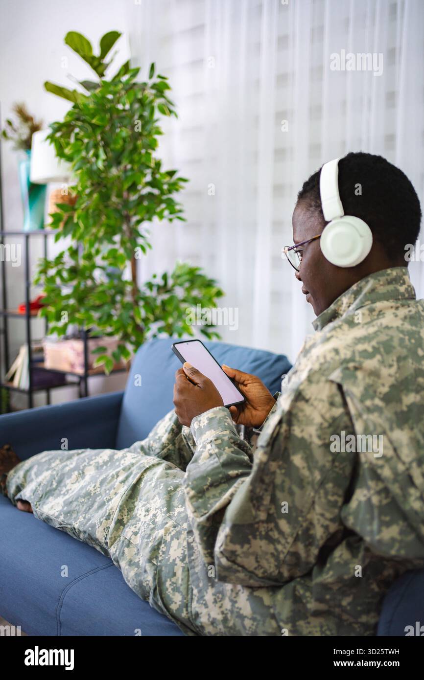 Soldat se relaxant sur le canapé à l'aide d'un smartphone et écoutant de la musique avec des écouteurs Banque D'Images