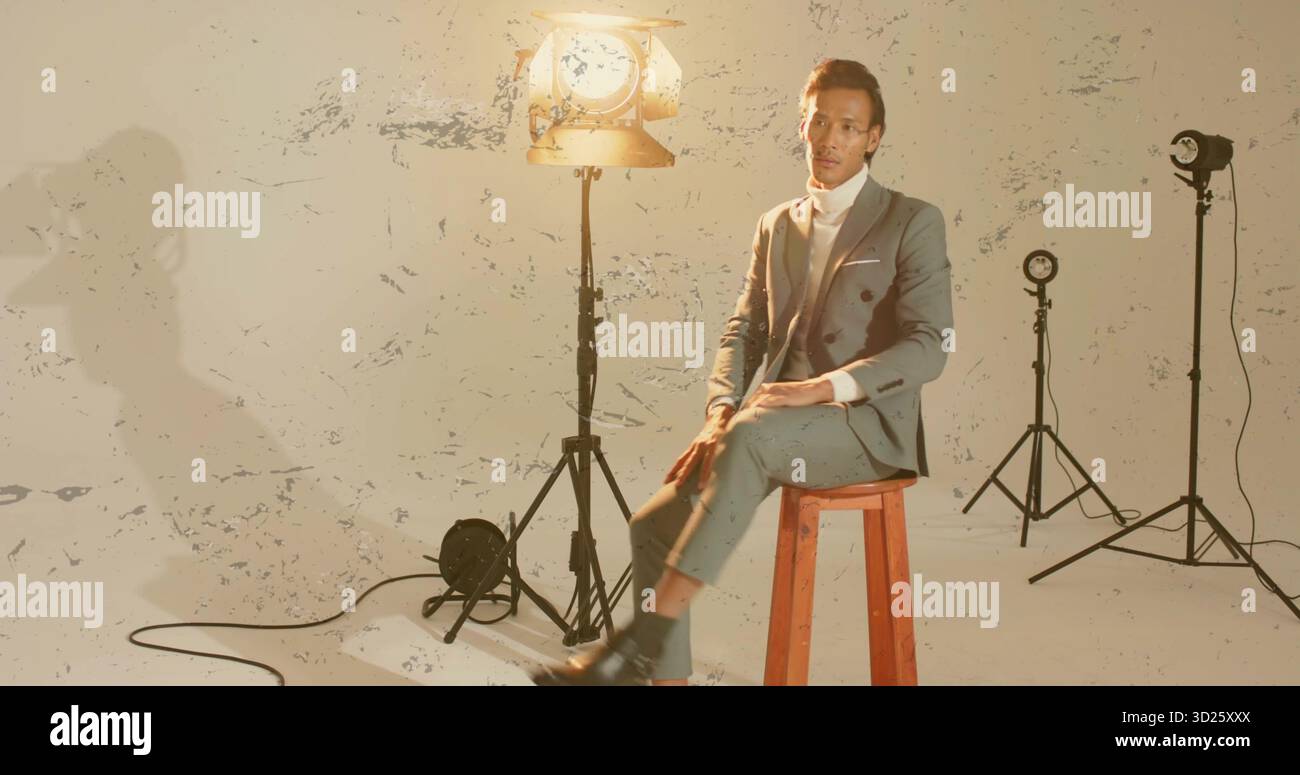 Homme asiatique portant un costume gris assis sur un tabouret dans le studio, avec des lumières, des cordes sur le mur peint-éclaboussé Banque D'Images