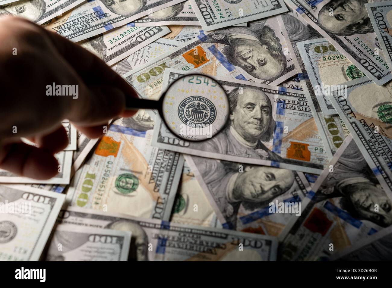 Une personne utilise une loupe pour inspecter de nombreux billets de centaines de dollars répartis sur une table, se concentrant intensément. Banque D'Images