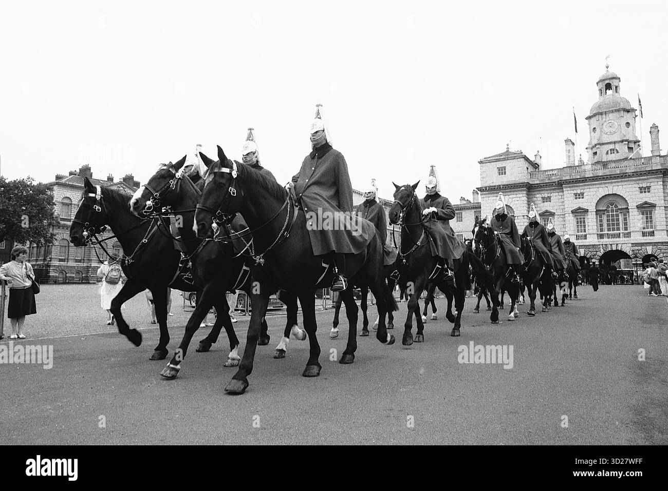 Une image en noir et blanc saisissante capture l'emblématique Horse Guards Parade à Londres. Les gardes montés, resplendissants en uniformes de cérémonie, foulent délibérément les pavés, un témoignage de siècles de tradition britannique. L'imposant bâtiment du Mémorial du duc de Wellington se dresse fièrement en arrière-plan, complétant une scène chargée d'histoire et de pageanterie. Banque D'Images