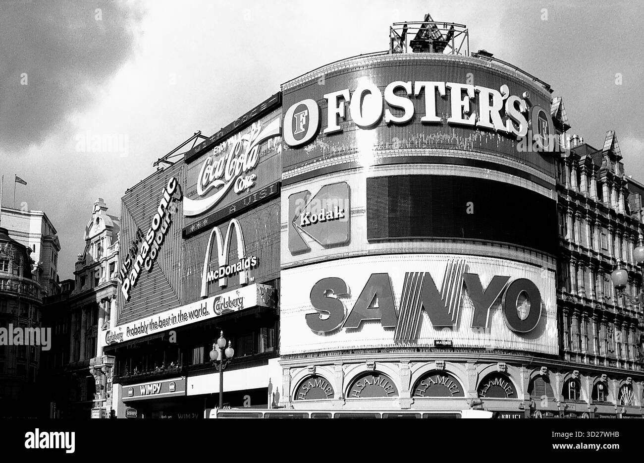 Une photographie en noir et blanc saisissante capture l'emblématique bâtiment de Forster à Londres. Les affichages publicitaires colossaux, mettant en vedette des marques comme Coca-Cola, Kodak et Sanyo, dominent l'horizon, mettant en valeur l'importance historique du bâtiment en tant que centre publicitaire majeur. Les détails architecturaux des bâtiments victoriens environnants ajoutent à l'attrait intemporel de l'image, créant une scène puissante du passé vibrant de Londres. Banque D'Images