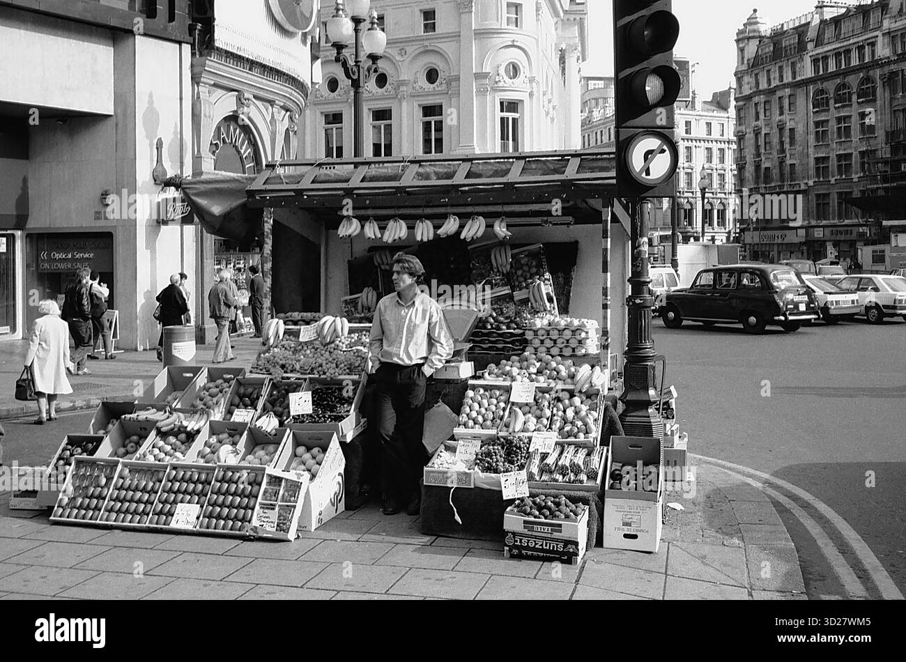 Une scène animée au cœur de Londres. Un étal dynamique de fruits et légumes occupe le premier plan, au milieu de l'architecture emblématique de Piccadilly Circus. La photographie classique en noir et blanc capture un moment emblématique de Londres, mettant en valeur la riche histoire et l'atmosphère animée de la ville. Banque D'Images