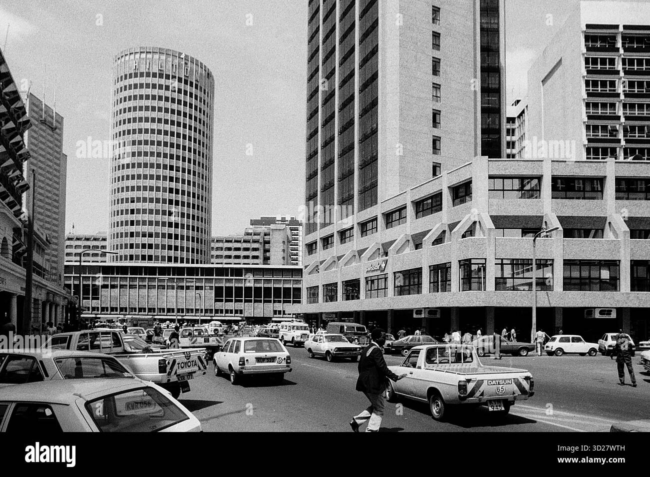 Quartier central des affaires de Nairobi, années 1970 Cette vue emblématique capture le cœur animé de la capitale du Kenya, dominé par l’imposant bâtiment de la Kenya International Bank et l’architecture moderne environnante. La scène de rue révèle un mélange vibrant de véhicules et de piétons, offrant un aperçu du développement rapide de la ville au cours des années 1970 Banque D'Images