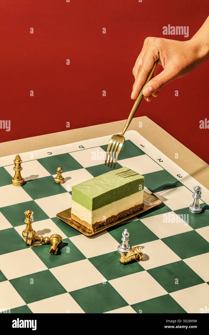 Scène d'échiquier surréaliste avec Cheesecake superposé Matcha percé par une fourchette dorée et des pièces tombées sur fond rouge Banque D'Images