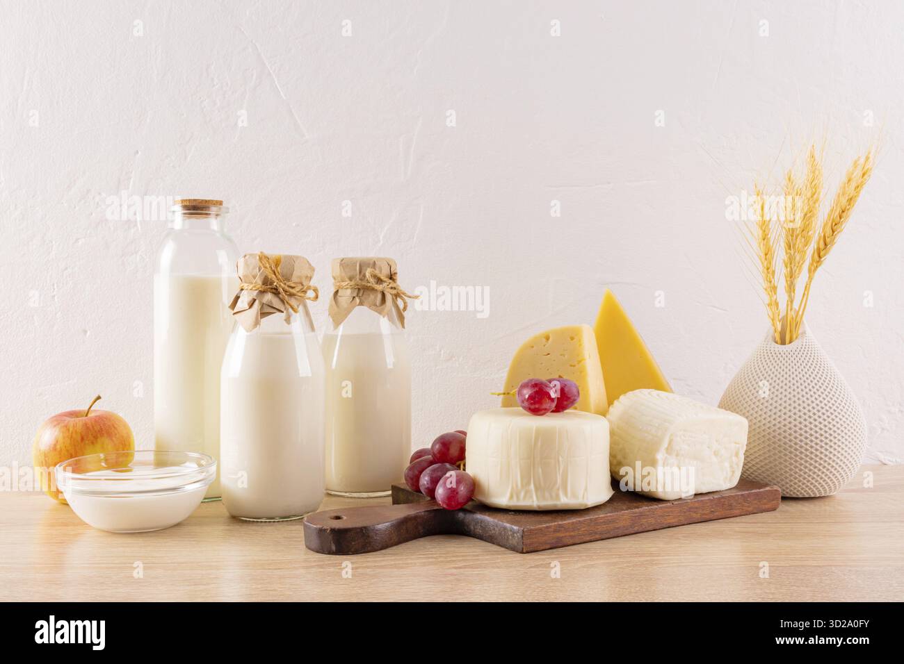 De nombreuses variétés de fromage sur une planche, des bouteilles de lait, du fromage cottage, de la crème sure, un bouquet d'épis de pain sur une table en bois. La fête de Shavuot Banque D'Images