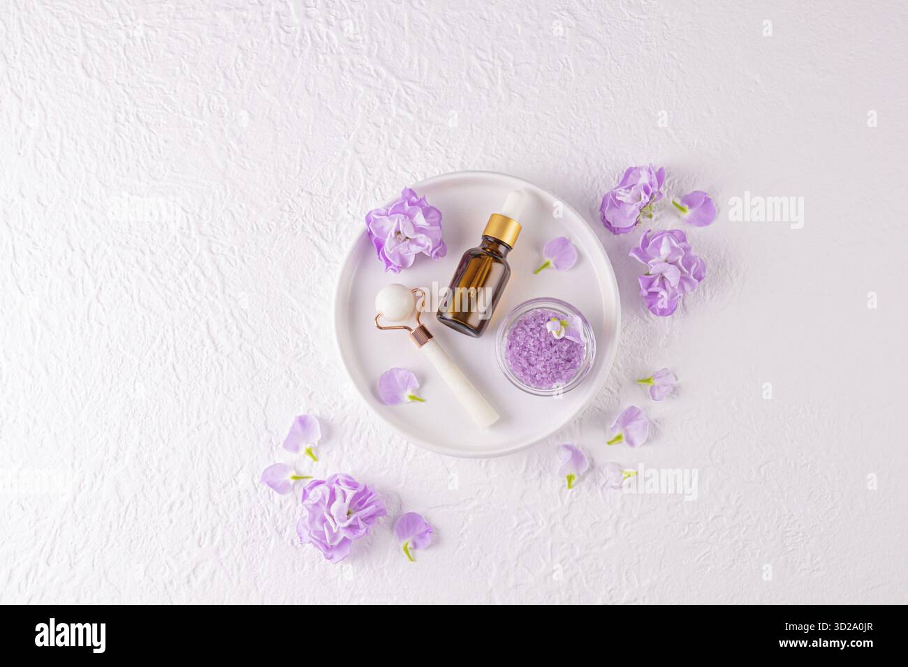 Set cosmétique pour spa et soins de la peau, massage. Rouleau de pierre, sels de bain, huile de massage sur plateau blanc en céramique. Vue de dessus. fleurs délicates lilas Banque D'Images