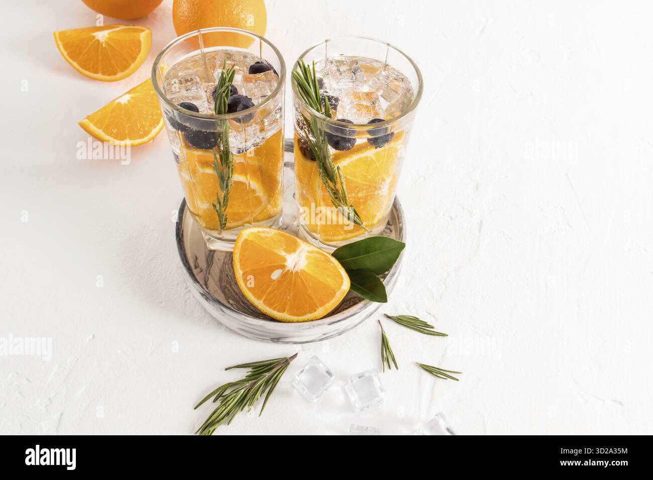 Deux verres avec de l'eau vitaminée avec des tranches d'orange, des bleuets, un brin de framboises, des glaçons sur une table blanche. une boisson rafraîchissante Banque D'Images