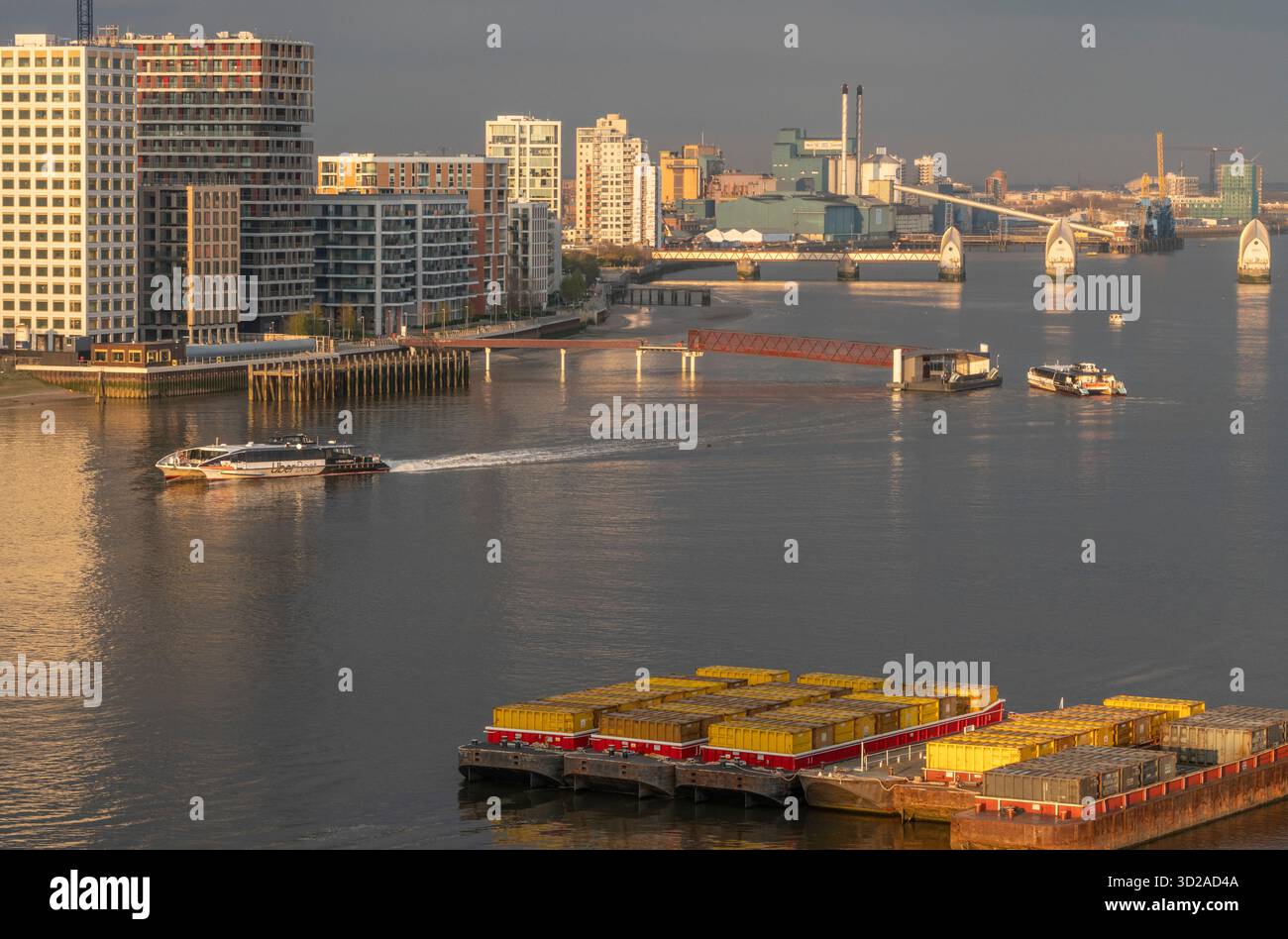Un bus fluvial Uber Boat Thames Clipper arrive à Royal Wharf Quay tandis qu'un autre part sur la rive nord de la Tamise avec les portes de contrôle des inondations de la barrière de la Tamise en arrière-plan à Londres, Royaume-Uni, le 15 avril 2023. Au premier plan se trouvent deux collecteurs passifs flottants de bois flotté (PDC) qui sont des cages pour collecter le bois flotté, les débris et les déchets plastiques de la rivière. Crédit : Rob Taggart/Alamy Banque D'Images