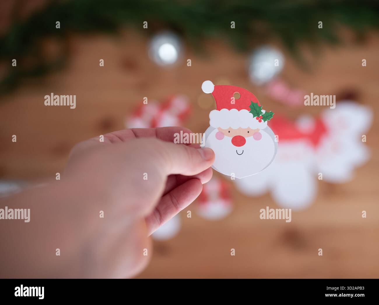 Une main festive tient une découpe en papier du Père Noël, entourée de décorations colorées des fêtes, créant une ambiance joyeuse et créative pour Christma Banque D'Images