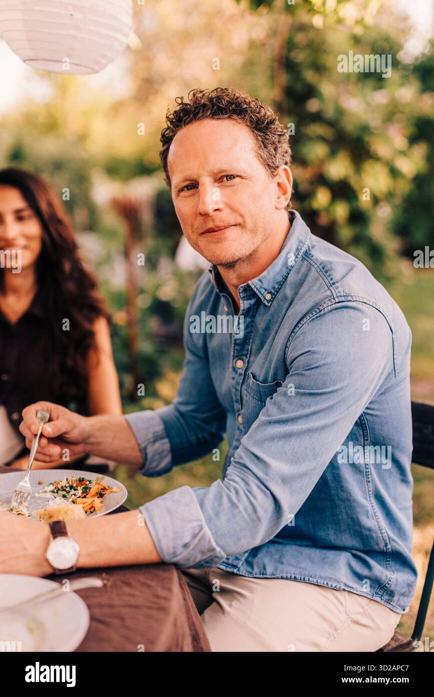 Portrait d'homme souriant mangeant de la nourriture tout en étant assis à la table à manger dans la cour arrière Banque D'Images